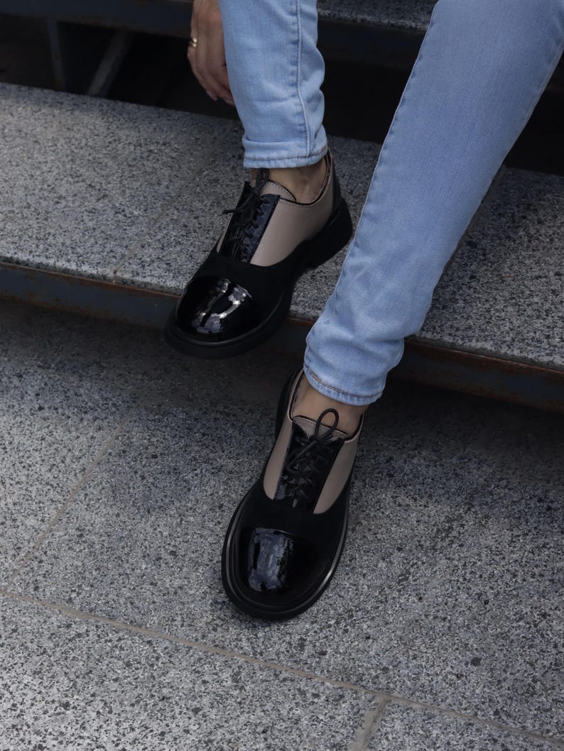 Puede incluir: Zapatos Oxford negros y beige con puntera de charol brillante y material similar al ante en la parte superior. Los zapatos tienen cordones negros y una suela gruesa negra. La persona lleva vaqueros azul claro.