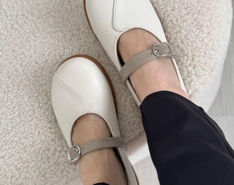 May include: White leather Mary Jane shoes with a light brown sole and a grey strap with a silver buckle. The shoes are on a white, fluffy surface. The person is wearing black trousers.