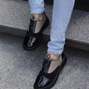 Puede incluir: Zapatos negros y beige estilo Oxford con puntera de charol brillante y un panel tipo ante. Los zapatos tienen cordones negros y una suela gruesa negra. La persona lleva vaqueros azul claro.