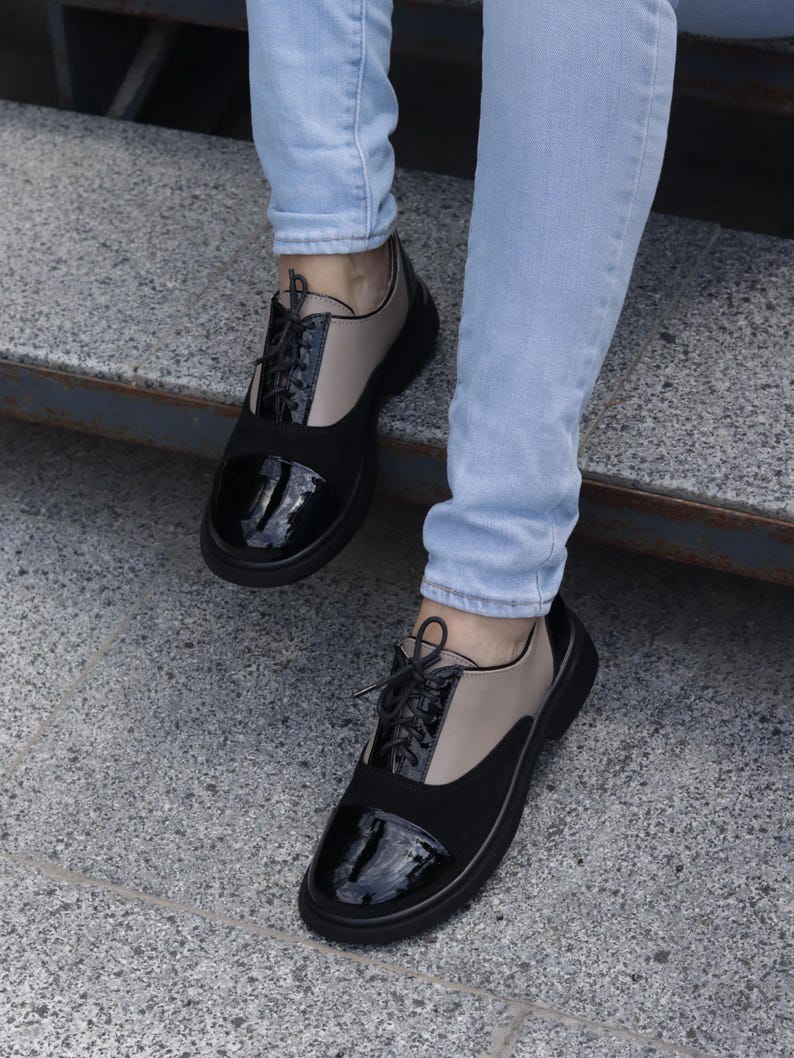 Puede incluir: Zapatos negros y beige estilo Oxford con puntera de charol brillante y un panel tipo ante. Los zapatos tienen cordones negros y una suela gruesa negra. La persona lleva vaqueros azul claro.
