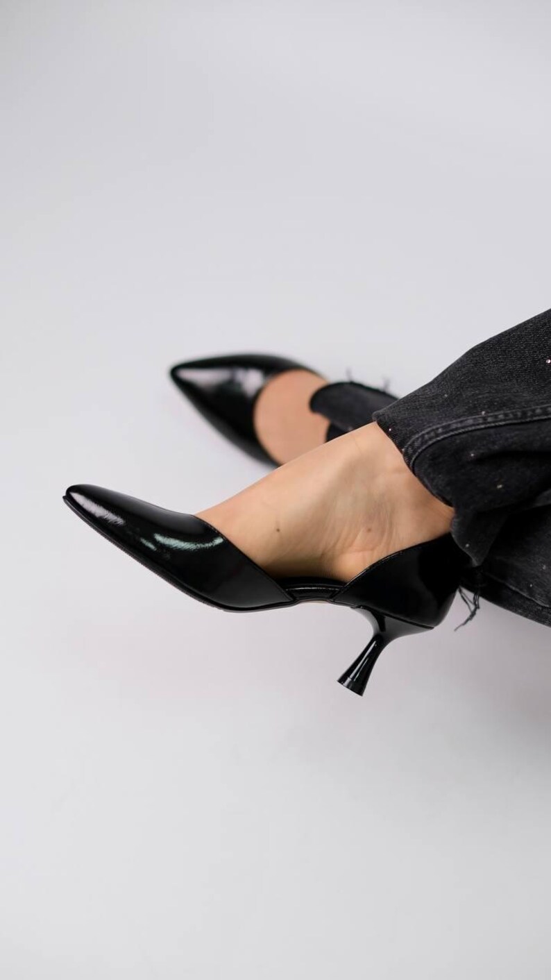 May include: A pair of black patent leather pumps featuring a pointed toe, a low kitten heel, and a side cutout design. The shoes are worn with dark gray denim jeans, showcasing a sleek and polished finish on the exterior of the footwear.