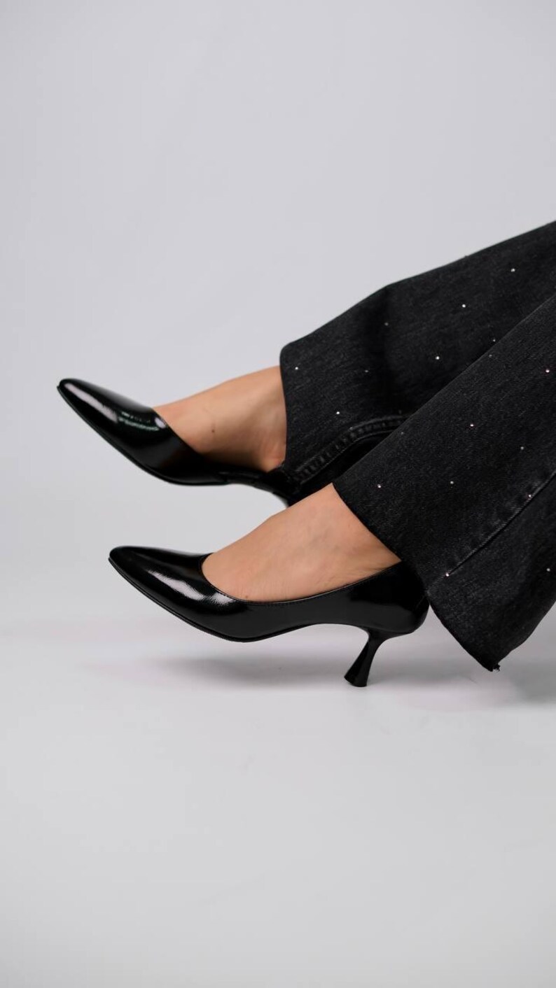 May include: Black patent leather pumps with a pointed toe and a low kitten heel, paired with black denim jeans featuring small, evenly spaced silver studs along the fabric. The shoes have a glossy finish and a simple, clean silhouette.