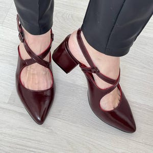 May include: A pair of burgundy heeled shoes with a criss-cross strap design. The shoes have a pointed toe and a block heel.
