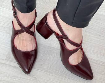 May include: A pair of burgundy heeled shoes with a criss-cross strap design. The shoes have a pointed toe and a block heel.