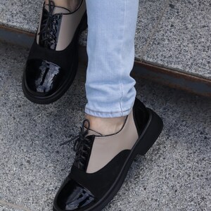Puede incluir: Zapatos Oxford bicolor con paneles negros, beige y negro brillante. Los zapatos tienen un dise&ntilde;o con cordones y una suela negra gruesa. La imagen muestra los zapatos desde abajo, con las piernas del usuario en vaqueros azul claro.