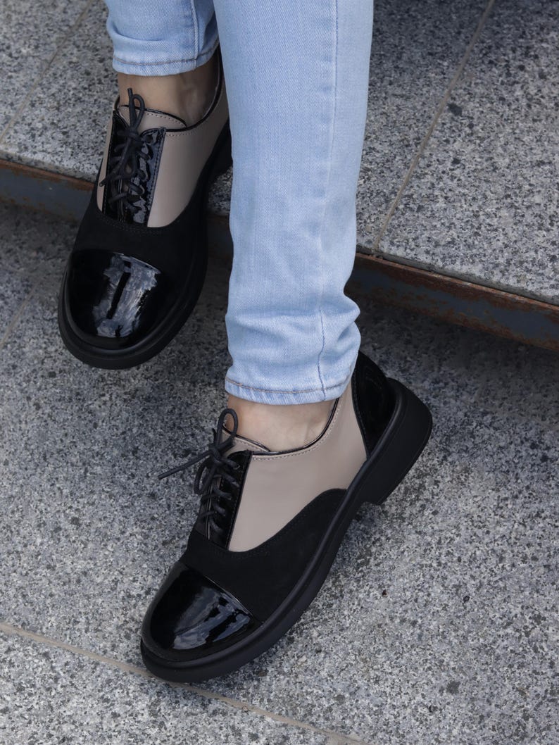 Puede incluir: Zapatos Oxford bicolor con paneles negros, beige y negro brillante. Los zapatos tienen un dise&ntilde;o con cordones y una suela negra gruesa. La imagen muestra los zapatos desde abajo, con las piernas del usuario en vaqueros azul claro.