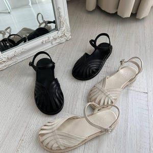 May include: Two pairs of flat sandals, one in black and one in cream, featuring a closed-toe design with multiple curved leather straps radiating from the center, an adjustable ankle strap with a metal buckle, and a flat sole for each shoe.