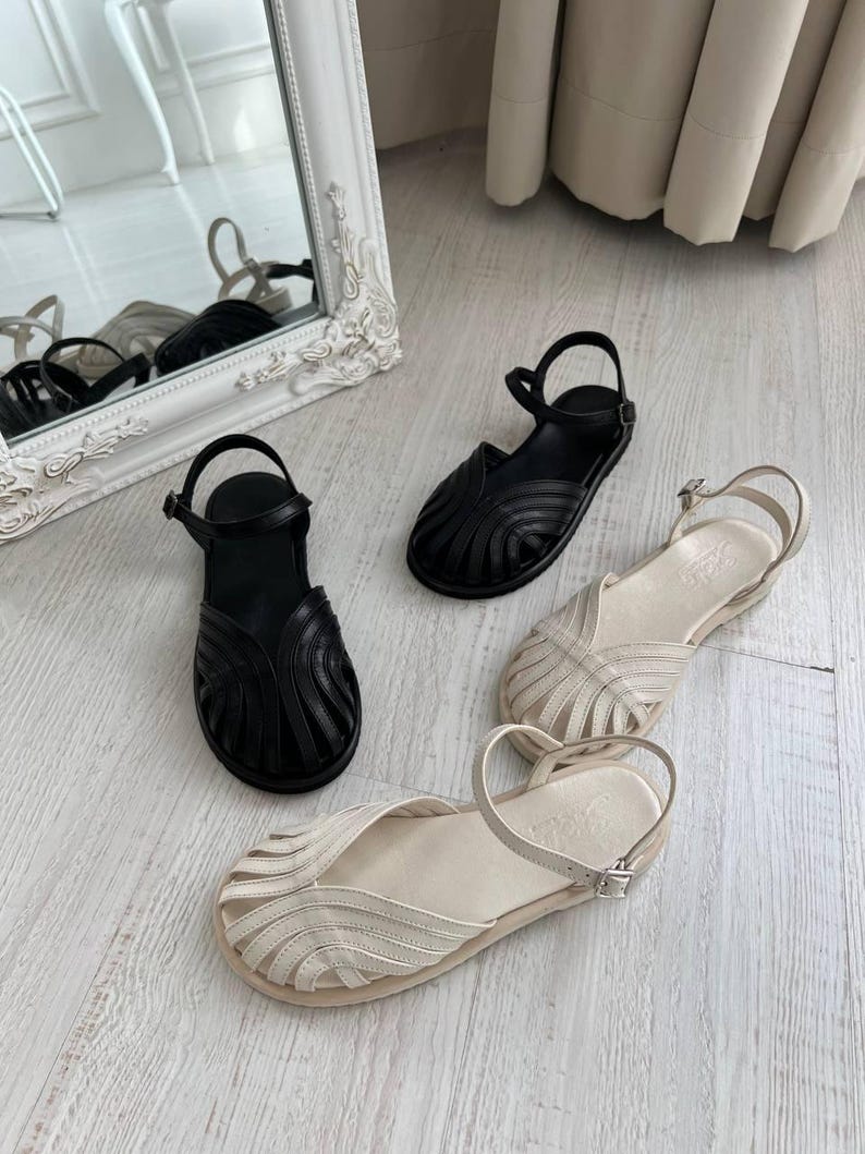 May include: Two pairs of flat sandals, one in black and one in cream, featuring a closed-toe design with multiple curved leather straps radiating from the center, an adjustable ankle strap with a metal buckle, and a flat sole for each shoe.