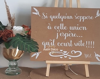 May include: A brown wooden sign on a small easel with white text that reads "Si quelqu'un s'oppose à cette union j'espère... qu'il court vite!!!!" A floral arrangement is in a gold-colored vase.