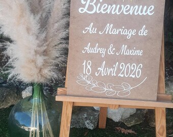 May include: A wooden easel displaying a wedding welcome sign. The sign is brown with white script, announcing the wedding of Audrey & Maxime on April 18, 2026. A large glass vase with pampas grass is to the left. The sign says "Bienvenue".