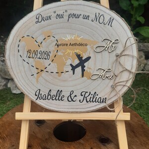 Könnte beinhalten: Ein hölzernes Hochzeitsschild auf einer kleinen Staffelei. Das Schild zeigt eine Weltkarte, ein Flugzeug und die Namen Isabelle & Kilian. Das Datum 12.09.2026 wird ebenfalls angezeigt. Der Text "Deux oui pour un NOM" steht oben.