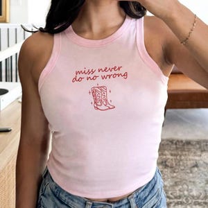May include: Pink tank top with a light pink trim and the text "miss never do no wrong" in red script. Below the text is a red illustration of cowboy boots. The top is made of a soft material.