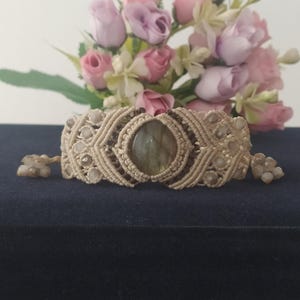 Pode incluir: Um bracelete de macramé bege com uma grande pedra de labradorita oval no centro. O bracelete é decorado com contas brancas menores.