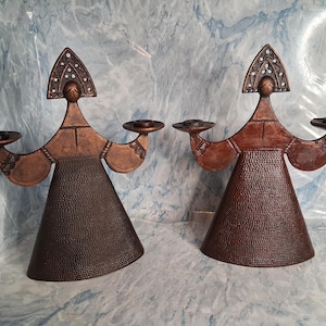 May include: Two bronze-colored candle holders with a unique design. Each holder features a figure with outstretched arms holding circular candle platforms. The figures have textured, cone-shaped bodies and ornate headpieces with white accents.