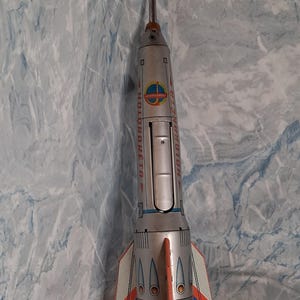 Pode incluir: Um foguete de brinquedo prateado vintage com detalhes em vermelho, branco e azul. O foguete tem um nariz longo e pontudo e barbatanas na base. O texto "HOLDRAMETA" está impresso na lateral. Um brinquedo retrô com tema de exploração espacial.