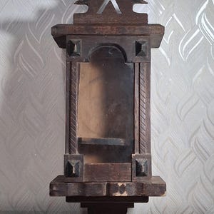 May include: A dark brown, antique wooden wall shelf with a decorative triangular top and a small shelf inside. The shelf has carved details and a distressed finish, suggesting age and use. The shelf is designed to be mounted on a wall.