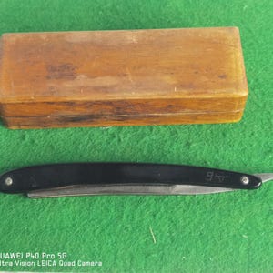 May include: A black straight razor with a silver blade and a curved handle, next to a rectangular wooden box. The razor has the letters "A B" etched on the blade. The items are on a green surface.