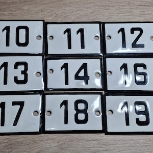 Könnte beinhalten: Neun rechteckige, weiße Emaille-Hausnummernschilder mit schwarzen Rändern. Die Zahlen 10, 11, 12, 13, 14, 16, 17, 18 und 19 sind in fetter schwarzer Schrift dargestellt. Jedes Schild hat zwei vorgebohrte Löcher.