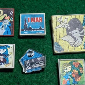 May include: A collection of vintage square pins with colorful illustrations. The pins feature various designs, including a building, a woman, and the text "1 MAY". The pins are set against a green background.