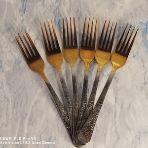 May include: A set of six gold and silver forks with ornate designs. The forks are arranged in a fan shape.