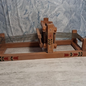 Könnte beinhalten: Ein hölzerner Garnwickler mit rechteckigem Rahmen und zentralem Mechanismus. Der Wickler ist mit roten und grünen Blumenmustern verziert. Garn ist zwischen den Seiten des Rahmens gespannt. Zum Halten von Garn zum Aufwickeln.