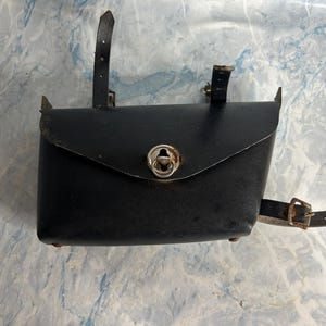 May include: A black leather pouch with a flap closure and a metal clasp. The pouch has two leather straps on the back and a buckle strap on the side. The pouch is approximately 15 cm wide and 10 cm tall.