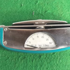 Könnte beinhalten: Vintage Messinstrument mit einem türkis-silbernen Gehäuse. Das Messgerät hat ein weißes Zifferblatt mit schwarzen Markierungen und dem Text "MADE IN USSR". Die Skala reicht von 0 bis 30. Das Instrument liegt auf einer grünen Oberfläche.