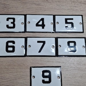 Könnte beinhalten: Eine Sammlung von weißen Emaille-Hausnummernschildern im Vintage-Stil mit schwarzen Ziffern. Die Nummern 3, 4, 5, 6, 7, 8 und 9 sind abgebildet. Jedes Schild hat einen schwarzen Rand und zwei Befestigungslöcher.