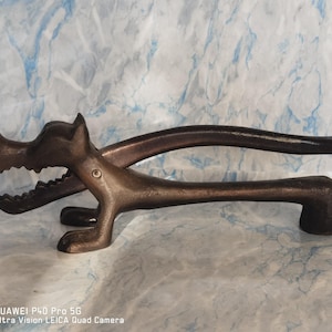 Può includere: Un schiaccianoci in ghisa a forma di cane con la bocca aperta e denti affilati. Lo schiaccianoci è marrone e ha un manico lungo e curvo.