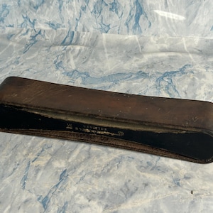 May include: A vintage, rectangular leather case with a curved shape. The case is a dark brown colour with visible wear and tear, suggesting age. The interior is black. Text is visible on the case.