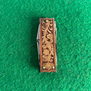 Puede incluir: Una navaja de bolsillo vintage con un mango decorativo. El mango presenta un diseño floral ornamentado en oro y marrón sobre un fondo oscuro, enmarcado por un borde rojo. Pequeños remaches plateados aseguran los paneles decorativos del mango.