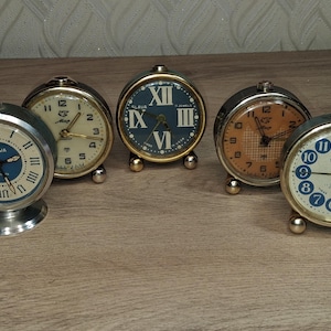 Puede incluir: Una colección de cinco despertadores antiguos. Los relojes tienen esferas redondas con números romanos y arábigos, en varios colores, incluyendo azul, blanco y naranja. Los relojes son de metal y tienen pequeños pies.