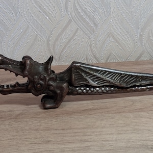 May include: A bronze-colored dragon-shaped nutcracker. The dragon's head is open, revealing teeth, and the wings are detailed with etched lines. The nutcracker rests on a wooden surface, showcasing its intricate design.