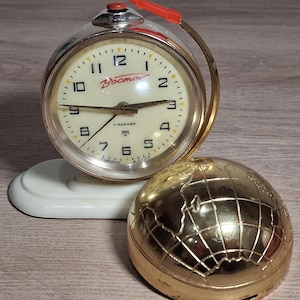 Puede incluir: Un reloj despertador vintage cromado con esfera crema, números negros y la palabra "Восток" en rojo. El reloj tiene un botón rojo y un asa dorada. Un objeto dorado en forma de globo se encuentra junto al reloj sobre una base blanca.