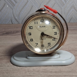 Könnte beinhalten: Ein goldfarbener Vintage-Wecker mit cremefarbenem Zifferblatt und schwarzen Ziffern. Die Uhr, mit der Aufschrift "SLAVA", hat einen roten Weckhebel und steht auf einem weißen Sockel. Das Zifferblatt zeigt die Uhrzeit 3:11 Uhr an.