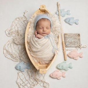 May include: A newborn baby wrapped in a cream-colored blanket and wearing a blue bonnet, nestled in a wooden boat. Decorative felt fish in blue and pink surround the boat, along with a fishing rod and a sign that reads "Gone Fishing."