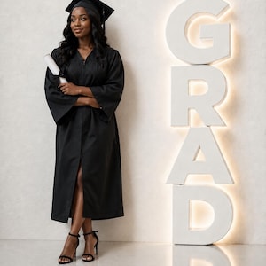 Op de afbeelding: Een persoon in een zwarte afstudeerjurk en baret, met een diploma. Het woord "GRAD" is in grote, verlichte witte letters gespeld. De persoon draagt zwarte sandalen met hoge hakken.