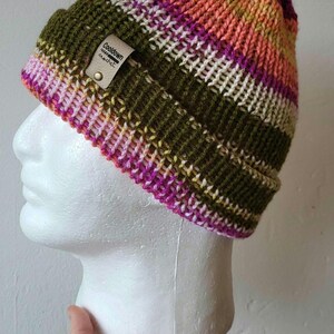 Puede incluir: Un gorro de punto colorido con rayas horizontales en verde oliva, rosa, blanco y naranja. El gorro tiene una pequeña etiqueta de cuero que dice "Cooldown Crochet". El gorro se muestra en una cabeza de maniquí blanca.