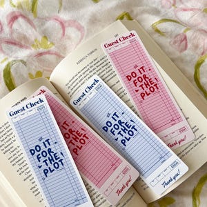 Peut inclure: Quatre marque-pages en forme de chèque d'hôtel rose et blanc avec du texte bleu qui dit "Do it for the plot". Les marque-pages sont sur une surface à motifs floraux rose et blanc.