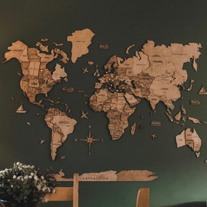 May include: A wooden world map with a compass and the words "Arctic Ocean", "Atlantic Ocean", "Indian Ocean", "Pacific Ocean", "Brazil", "Antarctica", "Russia", "China", "USA", "Canada", and "Greenland" printed on it. The map is mounted on a dark green wall. There are two wooden chairs and a wooden table in front of the map.