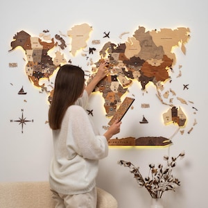 May include: A wooden world map with illuminated continents in various shades of brown and beige is mounted on a white wall. Decorative elements include a compass rose, airplanes, and sailboats. A person is holding a small wooden piece.