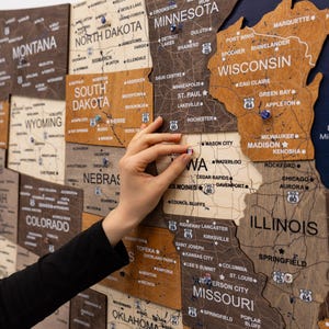May include: A multi-layered wooden wall map of the United States featuring laser-engraved state names, cities, and highway markers. The map is composed of various wood tones and includes small colorful push pins placed on specific locations.