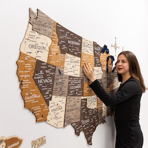 May include: A wooden United States map with state outlines and city names. The map is made of various wood tones and is mounted on a white wall. A person is touching the state of South Dakota. The map includes the Pacific Ocean and Hawaii.