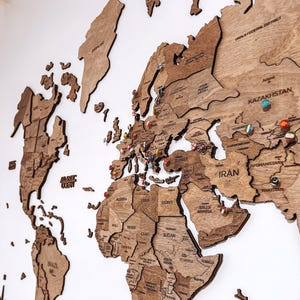 May include: A large wooden world map wall art piece featuring laser-cut continents and countries with engraved labels. The map is adorned with numerous small, colorful circular pins placed across various regions to mark specific locations.