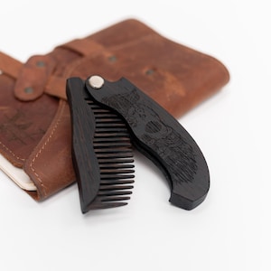 May include: A black folding comb with a detailed engraved design, likely wood, is partially open, revealing the comb's teeth. A brown leather case and notebook are in the background. The comb is designed for grooming.