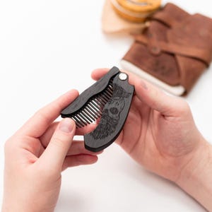 May include: A black wooden pocket comb with a skull and beard design. The comb is folded closed and has a metal hinge.