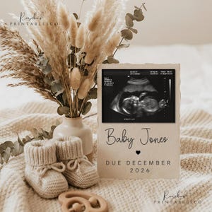 Könnte beinhalten: Eine beige Schwangerschaftsankündigung mit einem Ultraschallbild, dem Text "Baby Jones" und "Due December 2026". Das Schild befindet sich neben einem Paar gestrickter Babyschuhe, einem Holzspielzeug und einer Vase mit Trockenblumen.