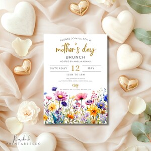 May include: A white invitation card with a floral design and gold lettering. The text reads "Please join us for a mother's day brunch hosted by Amelia Adams Saturday 12 May 11am to 1pm The Johnson's House Wildflower Lane, Daisy Hill RSVP to Amelia on by May 12th".