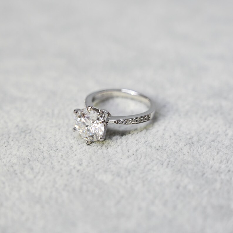 K&ouml;nnte beinhalten: Ein silberner Ring mit einem gro&szlig;en, klaren, runden Brillanten als Mittelst&uuml;ck und einer Reihe kleinerer Diamanten.