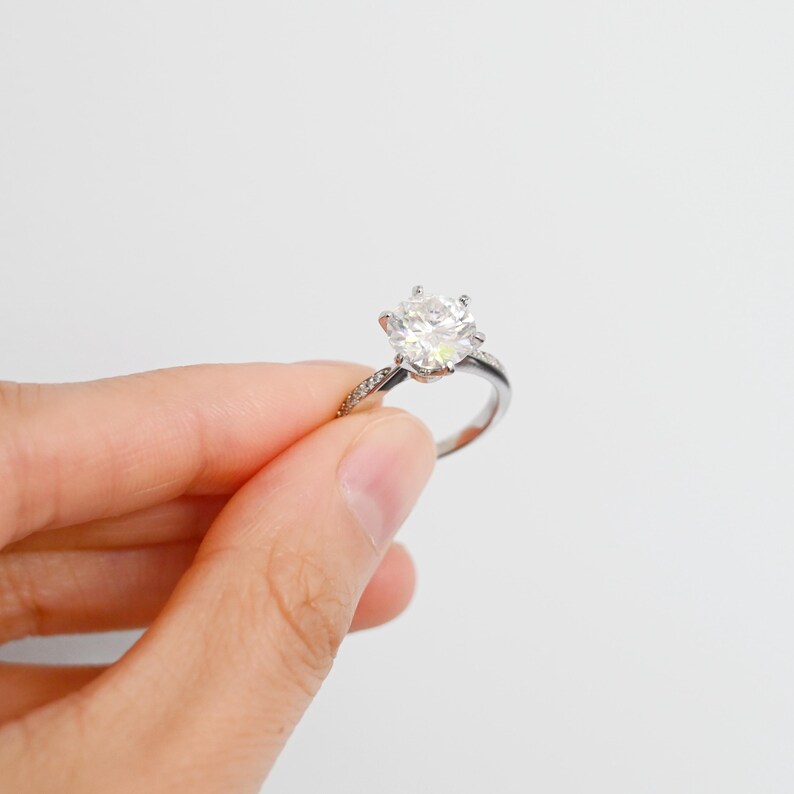 K&ouml;nnte beinhalten: Ein silberner Ring mit einem gro&szlig;en, klaren, runden Diamanten in der Mitte. Der Diamant ist von kleineren Diamanten umgeben.